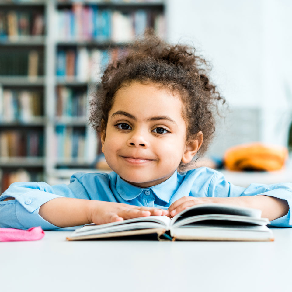 happy african american kid sitting and looking at camera near book and pink backpack