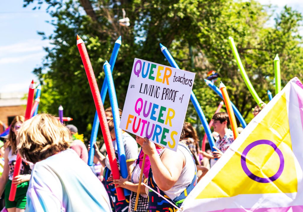 Utah Educators for Pride Group close up of sign "QUEER teachers are LIVING PROOF of QUEER FUTURES" in the LGBTQIA+ Pride Parade in Salt Lake City, Utah. June 04th, 2023 with multicolored pool noodle pencil props, and flags all unrecognizable.