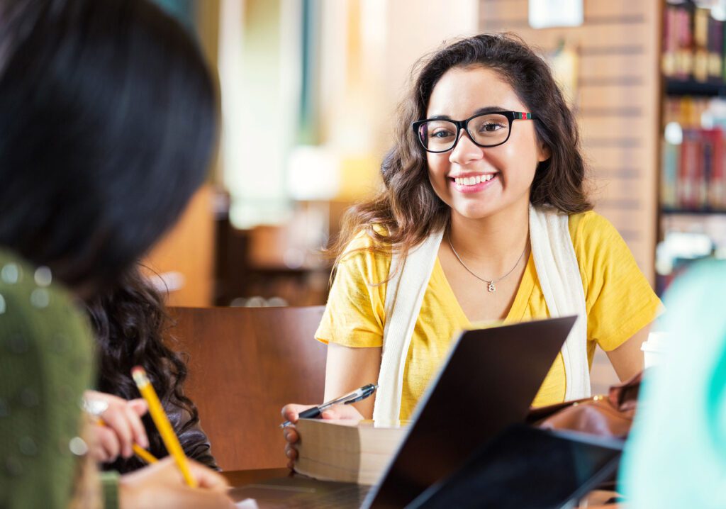 Trendy hipster Hispanic college age girl is smiling while sitting at tabe in library with group of friends. Students are working in study group to prepare for exam while using laptop computers, digital tablets, and other technology.