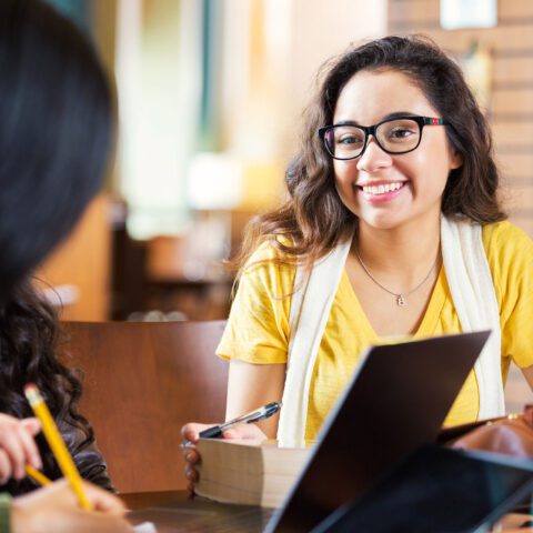 Trendy hipster Hispanic college age girl is smiling while sitting at tabe in library with group of friends. Students are working in study group to prepare for exam while using laptop computers, digital tablets, and other technology.